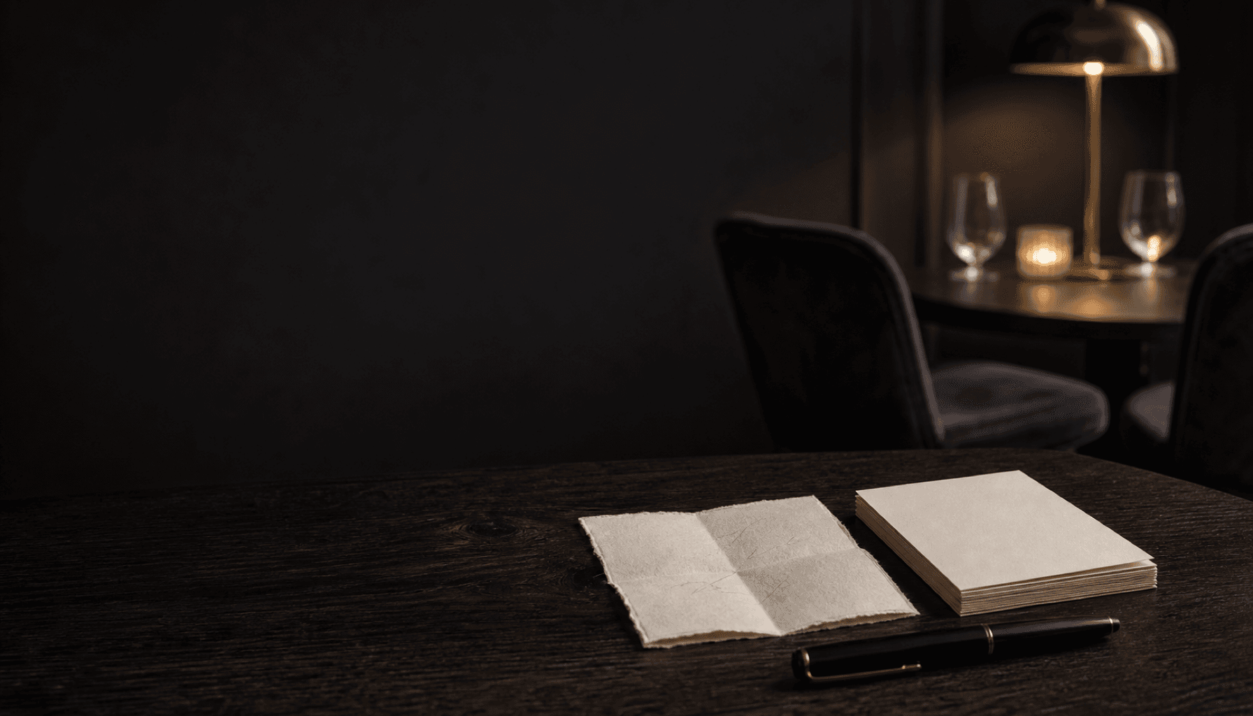 A folded handmade note and a neat stack of cream cards beside a fountain pen on a charcoal table in lamplight.