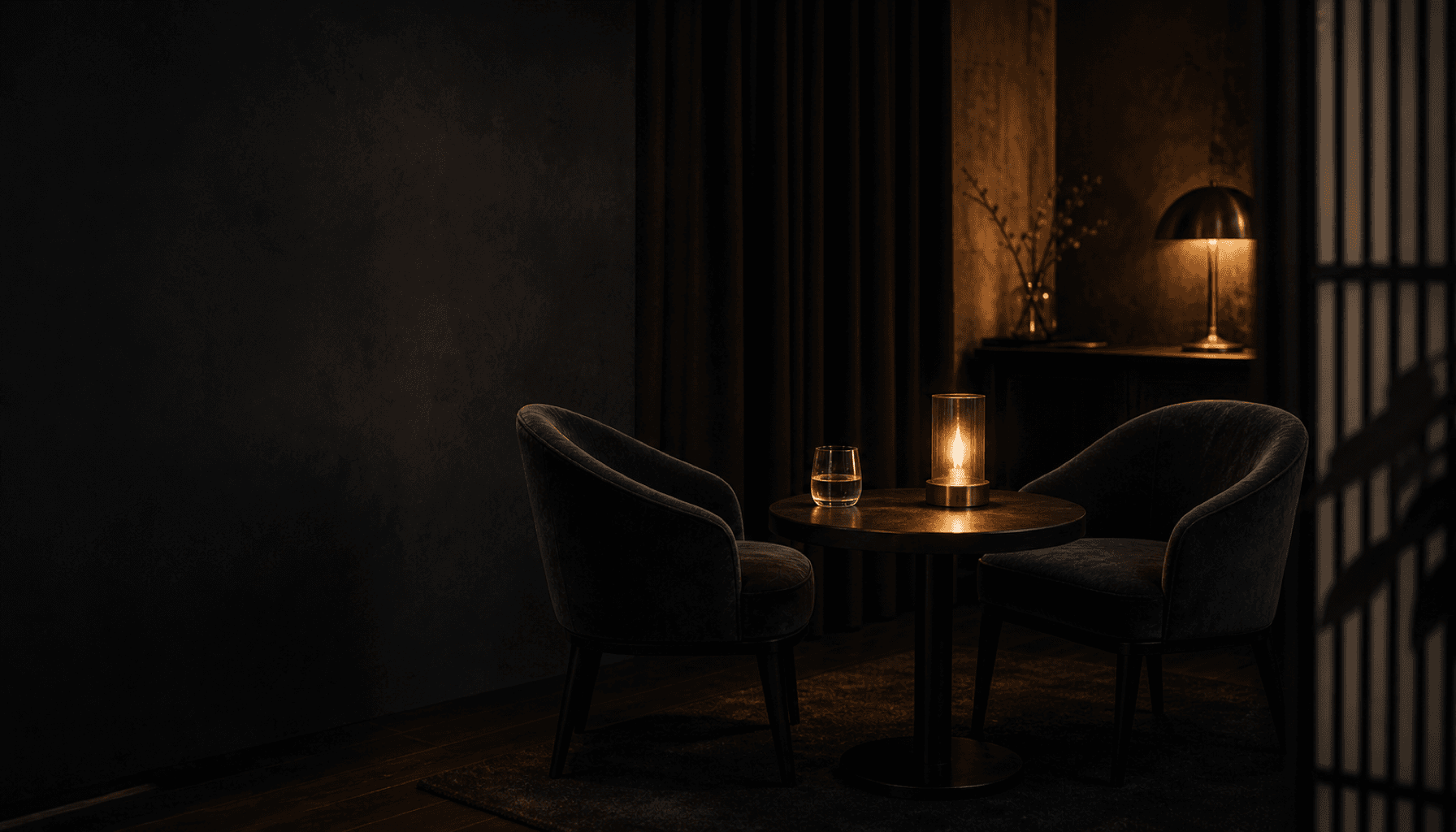 Two empty chairs drawn close at a small charcoal table, warm champagne light between them and a single glass set out.