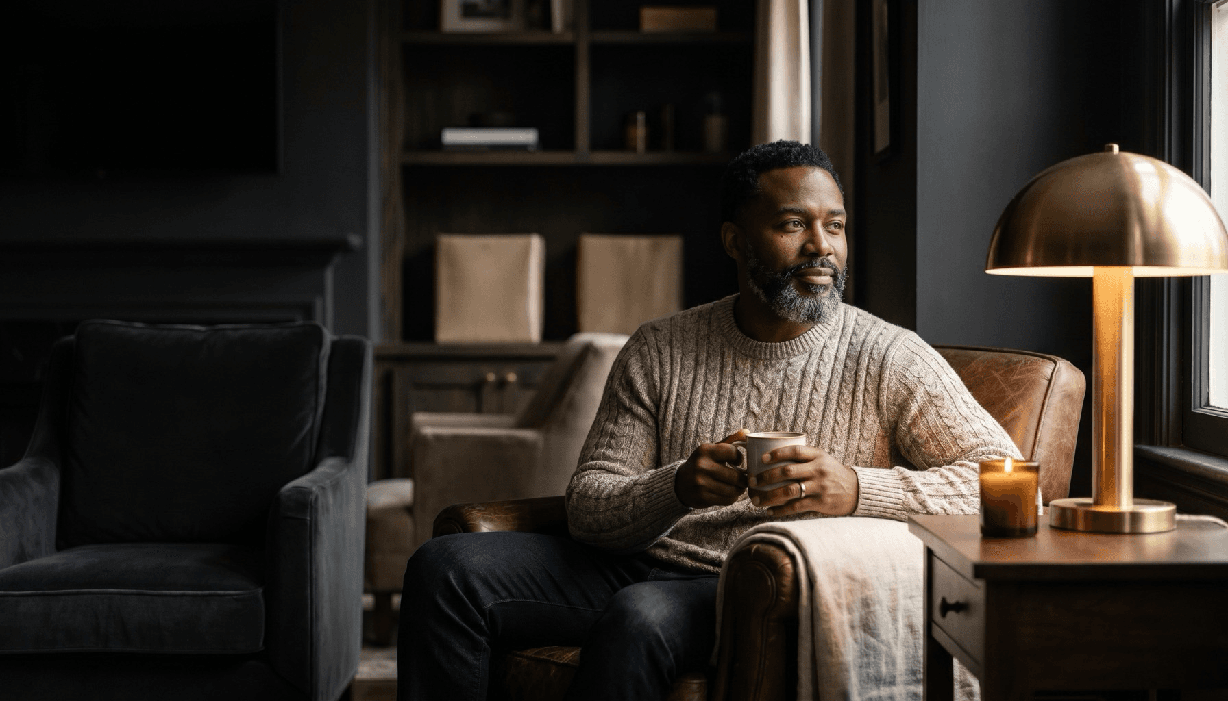 A composed man seated calmly with a cup of coffee in a warm, candlelit charcoal room.