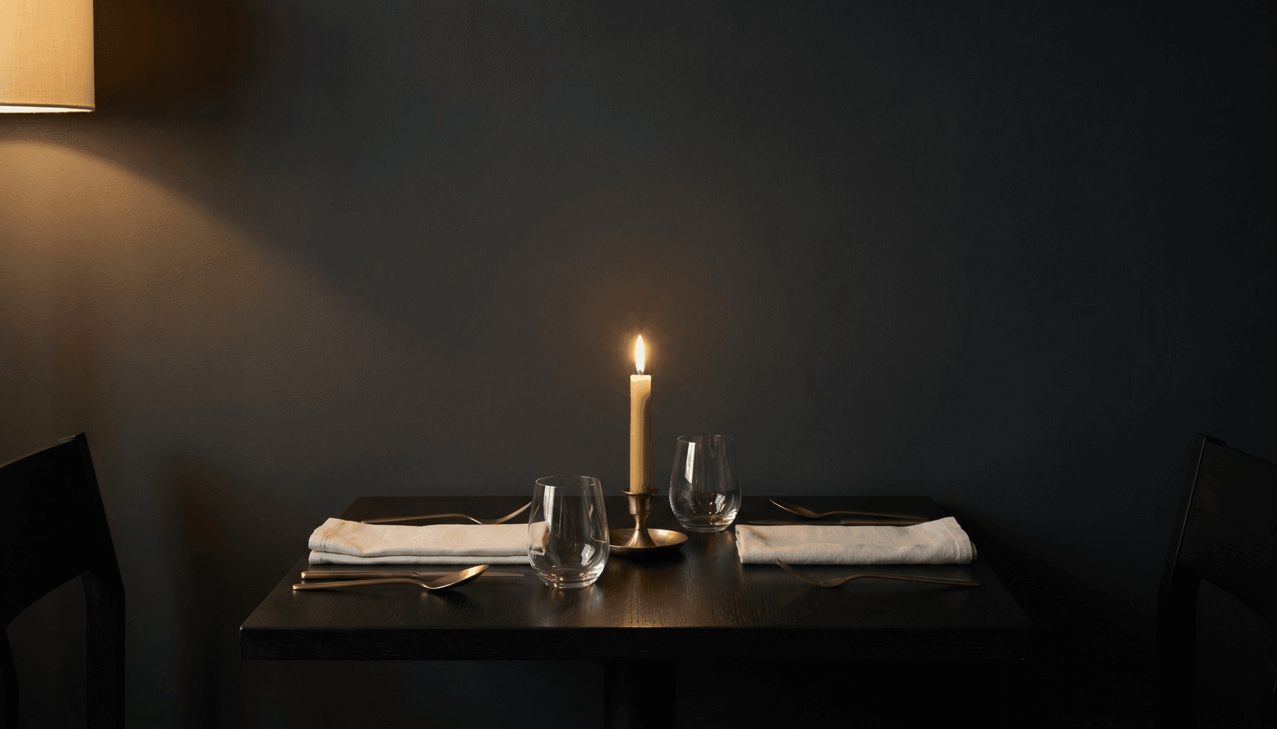 A single set table for two in a warm, candlelit charcoal room.
