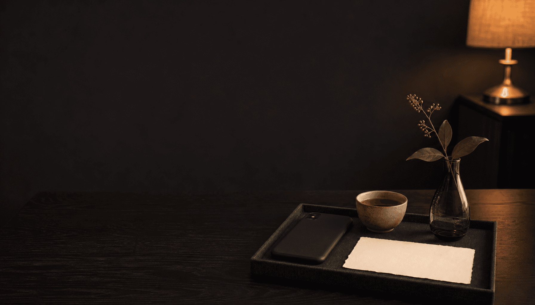 A face-down phone, a blank card, a cup of tea, and a single green stem on a tray in a lamplit charcoal room.