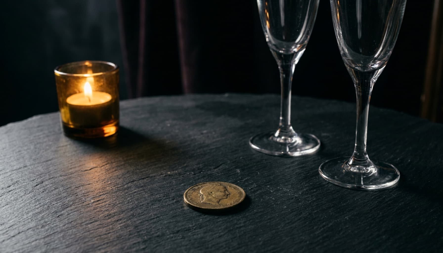 A brass coin beside two champagne glasses on a charcoal table in warm candlelight.
