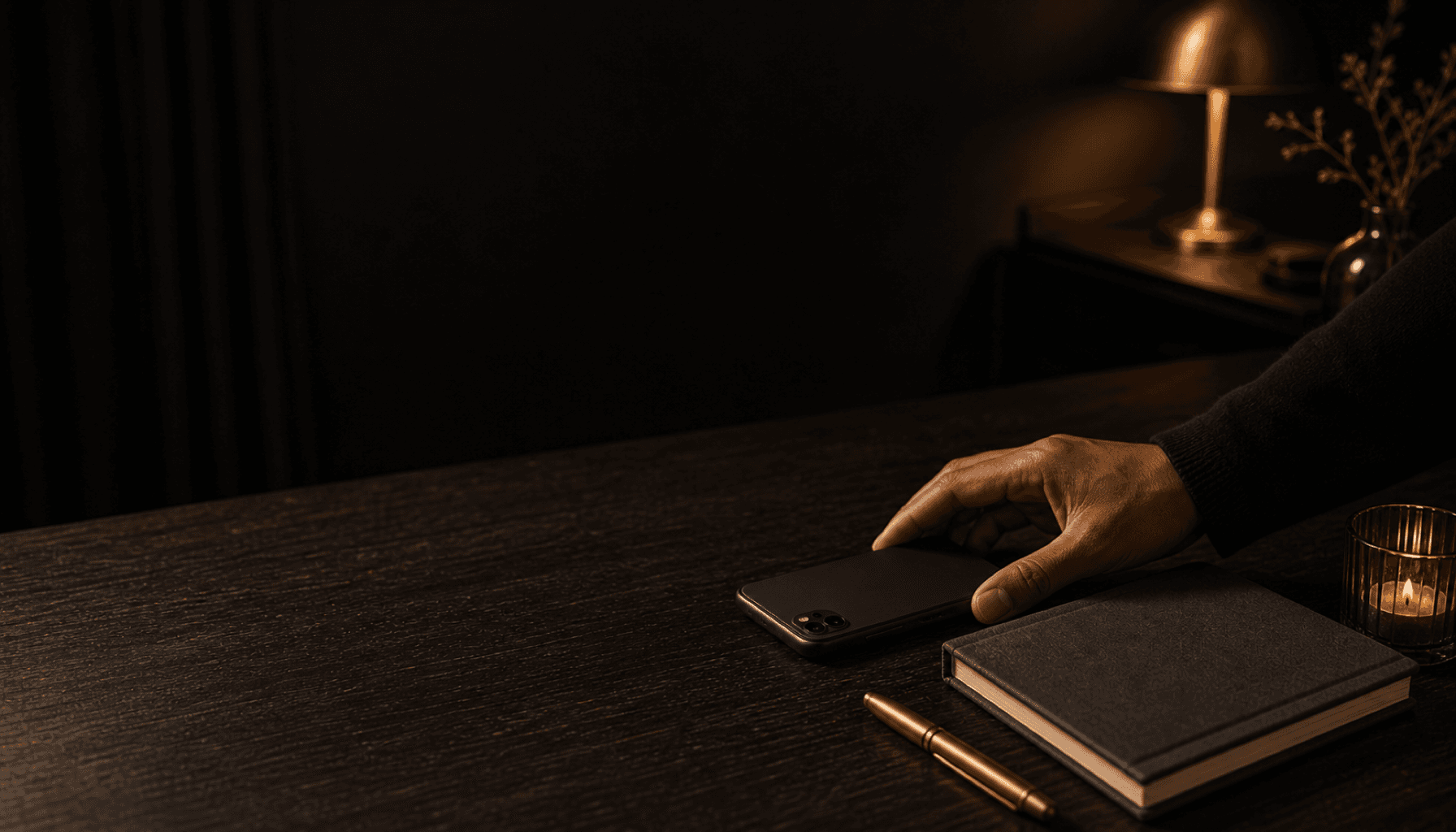 A hand placing a phone face-down beside a closed notebook on a lamplit charcoal table.